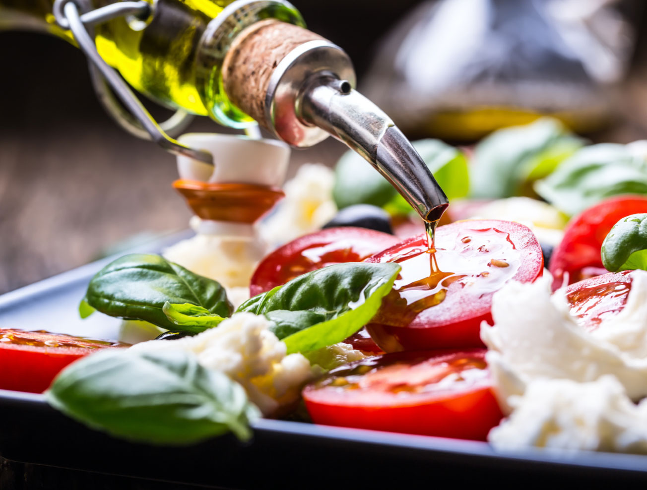 a bottle of olive oil being drizzled over tomatoes, cheese and basil