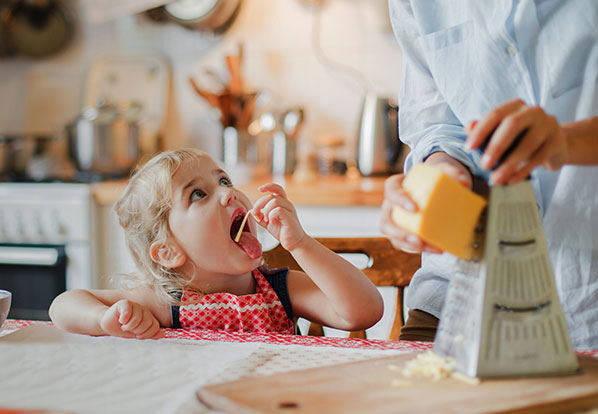a child eating a noodle while dad grates cheddar cheese